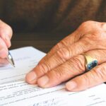 Close-up of a senior adult signing a legal document with a focus on hand and gold ring.