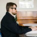 A visually impaired man reading Braille at a table in a warmly lit room with a piano in the background.