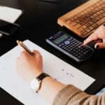 Close-up of hands working with a calculator and notebook on a desk, analyzing documents.